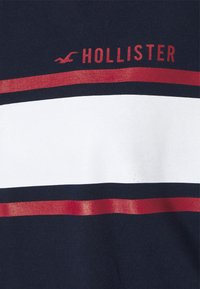 Granatowy materiał z dwiema poziomymi czerwonymi paskami i szerokim białym paskiem, z czerwonym napisem "Hollister" i logo mewy powyżej.