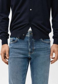 Mężczyzna ubrany w granatowy kardigan na białą koszulę i wyblakłe niebieskie dżinsy, stoi z ramionami luźno opuszczonymi wzdłuż ciała na jednolitym tle.