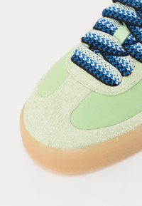 Främre delen av en ljusgrön mockasinsneaker med tjock sula i gummifärg och skosnören i blått, vitt och svart mönster.