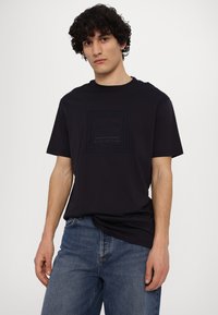 Camiseta de algodón negra con un logo cuadrado en relieve en azul marino oscuro y el texto "Armani Exchange Blue Edition". Mangas cortas, corte relajado.