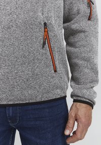 Jachetă fleece gri cu o suprafață texturată, având accente portocalii la fermoar și o margine neagră. De asemenea, este prezentată o mână odihnindu-se pe o pereche de blugi închiși la culoare.