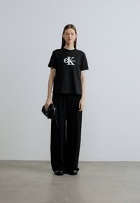 Svart bomulls-T-skjorte med hvit CK-logo, kombinert med vide svarte bukser. Modellen holder en liten svart veske, på en ensfarget bakgrunn.