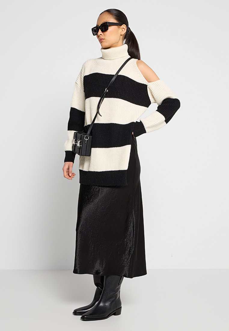 Zwart-witte gestreepte gebreide trui met een col en uitsnede op de schouder, gecombineerd met een glanzende zwarte midi-jurk en zwarte enkellaarsjes.