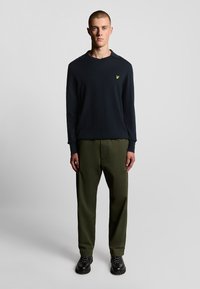 Camicia a maniche lunghe blu navy con scollo a V e logo giallo, abbinata a pantaloni verde oliva. Calzature nere completano l'outfit.