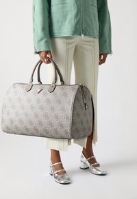 Sac de voyage à motifs gris avec texture lisse, poignées doubles, quincaillerie dorée et fermeture éclair, exposé à côté d'un pantalon vert pâle et de chaussures argentées.