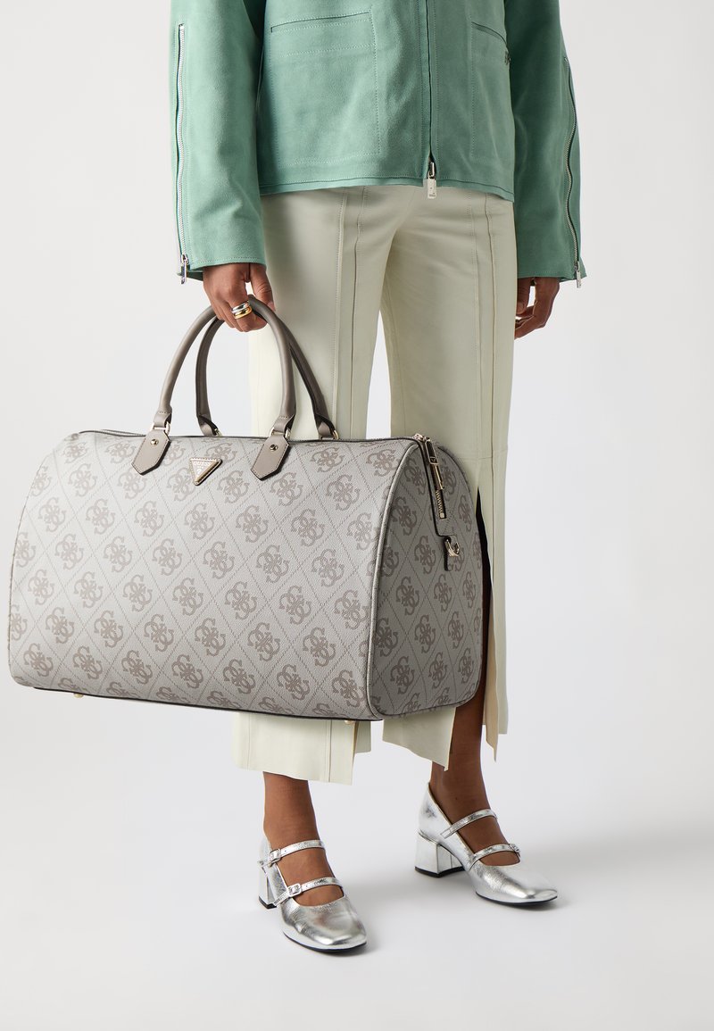Sac de voyage à motifs gris avec texture lisse, poignées doubles, quincaillerie dorée et fermeture éclair, exposé à côté d'un pantalon vert pâle et de chaussures argentées.