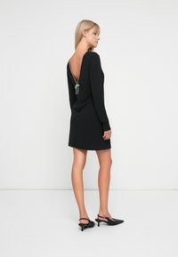 Kvinne står i en svart langermet minikjole med dyp V-rygg knyttet med en sølvsnor, iført svarte slingback heler.