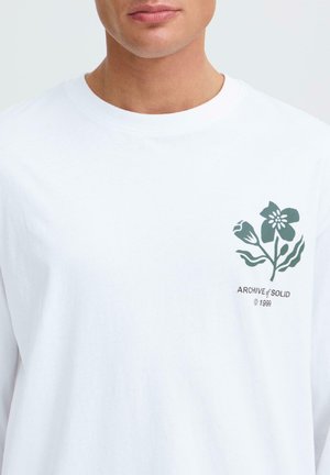 Hombre con camisa blanca de manga larga con una pequeña flor verde oscura y el texto "ARCHIVE of SOLID © 1999" en el pecho izquierdo.