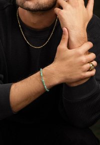 Gold chain necklace, dark textured sweater, turquoise beaded bracelet, large gold ring, visible hand tattoos, close-up of wrist and forearm.