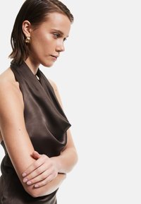 Asymmetrische bruine satijnen top met gedrapeerd stofdetail en een halternekkanaal. Model toont gekruiste armen en draagt ronde gouden oorbellen.