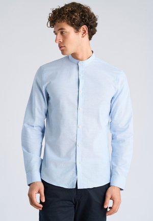 Giovane con capelli ricci che indossa una camicia celeste con colletto alla coreana e pantaloni scuri, che guarda verso sinistra su uno sfondo semplice.