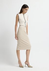 Ärmelloses, enges Oberteil in Creme mit schwarzem Besatz, kombiniert mit einem knielangen beigen Rock. Modell trägt metallische High Heels und posiert vor einem hellen Hintergrund.