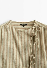 Blouse à rayures verticales beige et crème avec un col rond et trois nœuds en tissu attachés sur l'épaule droite.