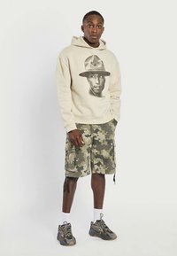 Hoodie beige arborant un graphisme du visage d'une personne, associé à un short cargo camouflage. Le modèle porte des chaussettes blanches et des baskets épaisses.