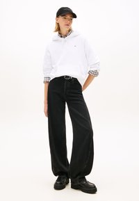 Sweat à capuche blanc avec une poche frontale, superposé à une chemise à carreaux, associé à un jean large noir et des chaussures à plateforme noires. Casquette noire en accessoire.