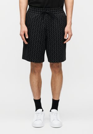 Homme portant un short à cordon de serrage noir à motifs, des chaussettes noires et des baskets blanches, debout devant un fond clair uni.