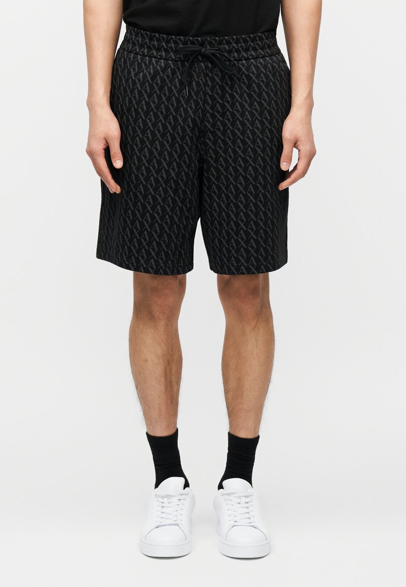 Homme portant un short à cordon de serrage noir à motifs, des chaussettes noires et des baskets blanches, debout devant un fond clair uni.