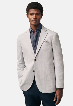 Blazer gris clair avec une texture en lin, comportant deux boutons, un col cranté et un pochette à motifs. Porté par-dessus une chemise sombre.