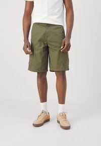 Pantalones cortos de cargo color verde oliva con dos bolsillos laterales, fabricados en una mezcla de algodón. Combinados con zapatillas de ante beige y calcetines blancos.