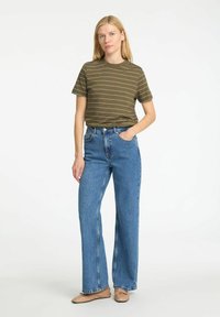T-shirt a righe color verde oliva con colletto rotondo, abbinata a jeans a vita alta di colore azzurro chiaro a gamba larga e scarpe basse di colore chiaro.