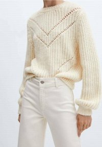 Pull en tricot couleur crème avec un motif texturé, présentant des détails en chevron ajourés. Associé à un jean blanc taille haute.