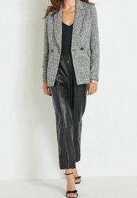 Blazer grigio in tweed con rever a la moda e due bottoni, abbinato a pantaloni neri in finta pelle a gamba affusolata e tacchi neri con cinturini.