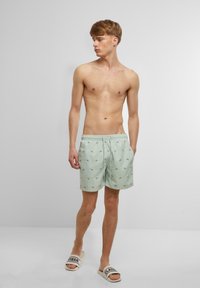 Svijetlozelene kupaće gaće s plavim uzorkom riba, elastičnom strukom, dva bočna džepa i opuštenim krojem; uparene sa svijetlim kliznim sandalama.