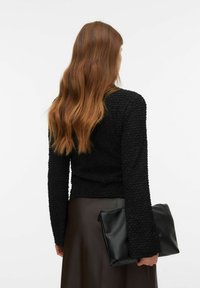 Vrouw met lang roodachtig haar in een gestructureerde zwarte top en een donkere rok houdt een grote zwarte clutch vast tegen een witte achtergrond.