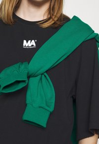 Person iført en sort Martin Asbjørn T-shirt med en grøn sweatshirt bundet over skulderen, der kun viser den øverste del af torsoen.