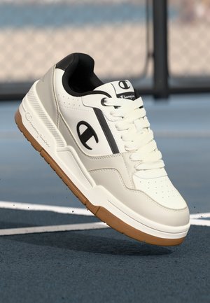White leather sneaker with black accents, featuring perforated toe, white laces, and a gum sole. Brand logo displayed on the side.