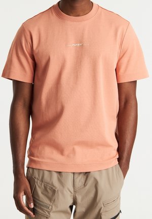 Homme portant un t-shirt à manches courtes col rond saumon clair et un pantalon cargo beige avec poches, debout avec une main dans la poche.