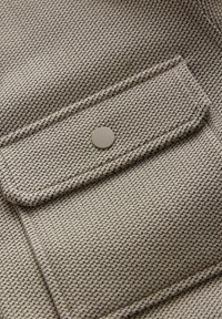 Beige mønster stof med en tekstureret overflade, der har en flaplomme og en skinnende metal snapknap i midten.