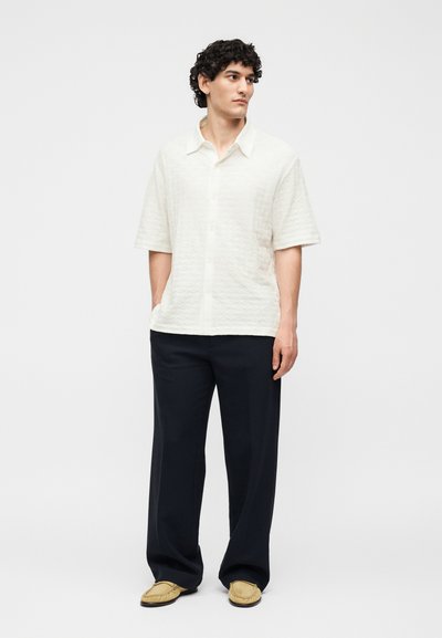 Νεαρός άνδρας στέκεται με τα χέρια στις τσέπες φορώντας μια λευκή υφασμάτινη κοντομάνικη πουκαμίσα, μαύρο φαρδύ παντελόνι και μπεζ μοκασίνια.