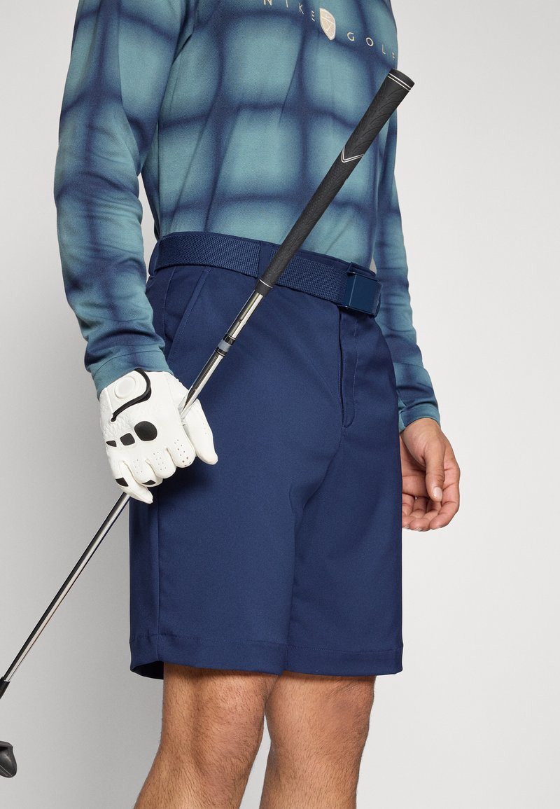 Marineblaue Golfshorts mit glatter Textur, ausgestattet mit einem Gürtel und einem sauberen Saum. Weißer Golfhandschuh mit schwarzem Akzent, der einen Golfschläger hält.