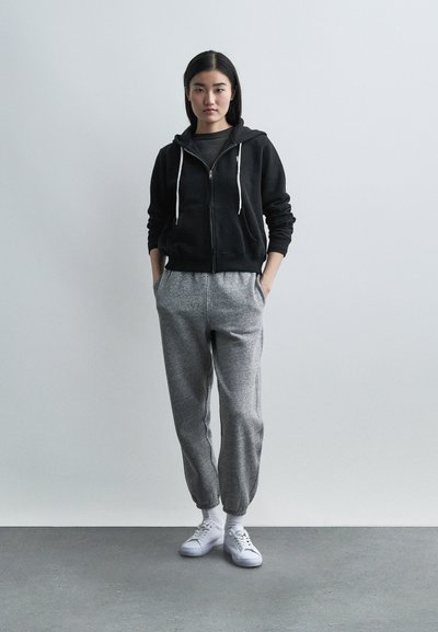 Νέα γυναίκα που στέκεται με τα χέρια στις τσέπες, φοράει μαύρο φούτερ με φερμουάρ, γκρίζο φόρμα παντελόνι, λευκές κάλτσες και λευκά αθλητικά παπούτσια σε γκρίζο πάτωμα.