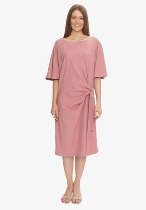 Kobieta stoi i się uśmiecha, nosi pudrowo-różową sukienkę do kolan z szerokimi rękawami i węzłem z boku, beżowe sandały na obcasie z odkrytymi palcami.