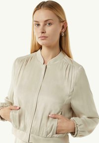 Beżowa kurtka bomberka z kołnierzem mandarynkowym, plisowanymi ramionami i elastycznymi mankietami. Posiada boczne kieszenie i gładką, błyszczącą fakturę.