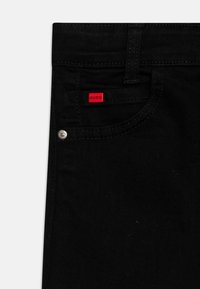 Vaqueros de mezclilla negra con un bolsillo delantero que presenta una pequeña etiqueta roja de HUGO, detalle de herrajes plateados y una textura sutil.