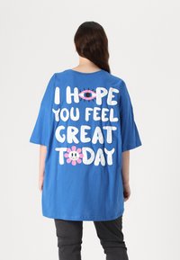 Blue oversized t-shirt featuring large white text: "I HOPE YOU FEEL GREAT TODAY," with a pink flower and smiley graphics. Short sleeves.