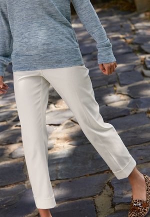 Pantalon blanc sur mesure avec ourlet retroussé, associé à un pull bleu texturé. Des chaussures marron à imprimé léopard complètent la tenue.