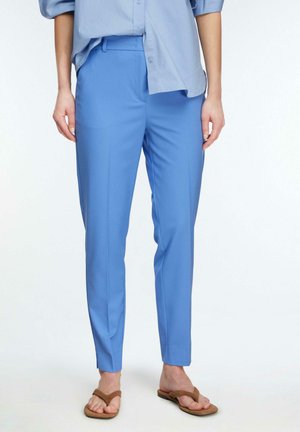 Osoba ubrana w jasnoniebieskie, dopasowane spodnie, pasującą, niezałożoną koszulę oraz brązowe, płaskie klapki, stojąca na tle białego tła.