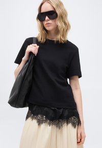 Γυναίκα με σγουρά ξανθά μαλλιά που φοράει μεγάλα μαύρα γυαλιά ηλίου, μαύρο μπλουζάκι, μπεζ φούστα με μαύρη δαντέλα και κρατά μια μαύρη τσάντα.