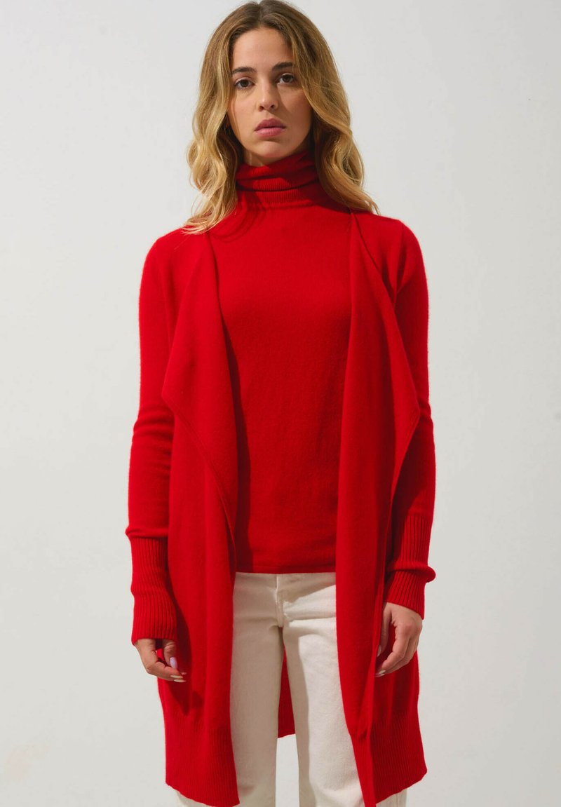 Roter, gestrickter langer Cardigan mit offenem Vorderteil, gerippten Bündchen und einem Rollkragenpullover darunter; getragen mit cremefarbenen Hosen.
