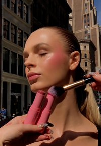 Two pink cream blush sticks held near a model's cheek, applying makeup with a brush. Model has smooth skin and a natural glow.