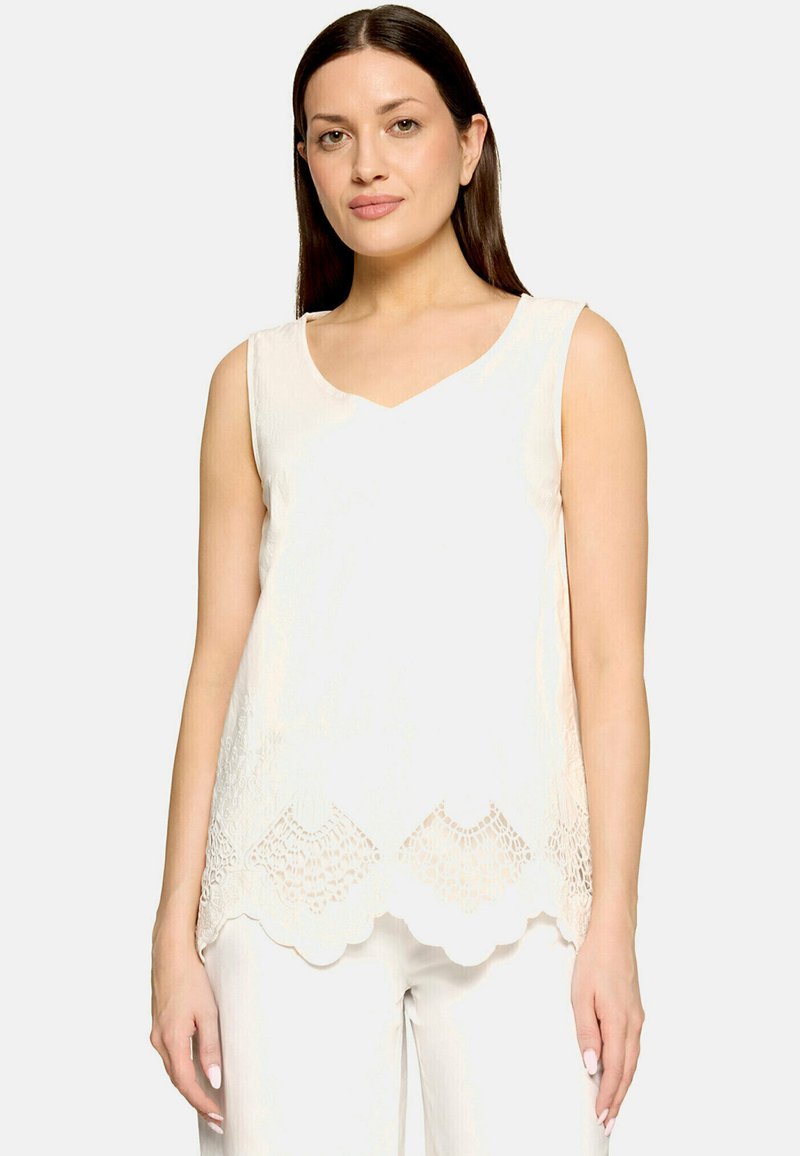 Femme aux cheveux raides et foncés, vêtue d'un haut blanc sans manches avec un ourlet en dentelle et d'un pantalon blanc, debout devant un fond clair uni.