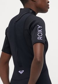 Juodasis neopreno trumpomis rankovėmis wetsuit'as su purpuriniu "ROXY" prekės ženklu ant rankos ir širdies logotipo akcentu apatinėje nugaros dalyje.