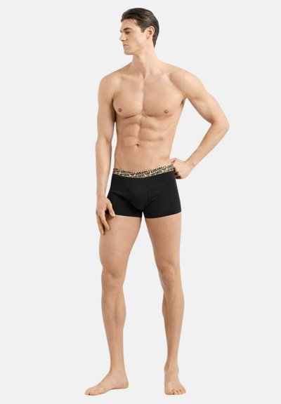 Uomo muscoloso in piedi a piedi nudi, che indossa boxer neri con una fascia in vita dorata decorata, guarda verso sinistra con una mano sui fianchi.