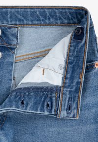 Jeans in denim con un lavaggio blu medio, caratterizzati da cuciture color tan, bottoni in metallo e una fodera interna bianca con chiusure a bottone.