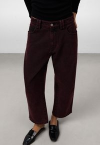 Pantaloni a zampa d'elefante in denim bordeaux con vita alta, tasche anteriori e cuciture visibili. Abbinati a scarpe nere senza lacci.
