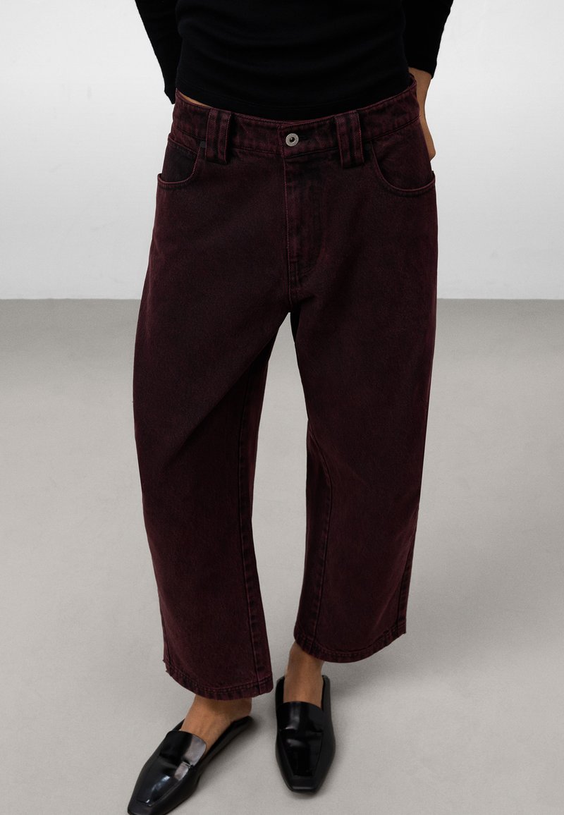 Pantaloni a zampa d'elefante in denim bordeaux con vita alta, tasche anteriori e cuciture visibili. Abbinati a scarpe nere senza lacci.